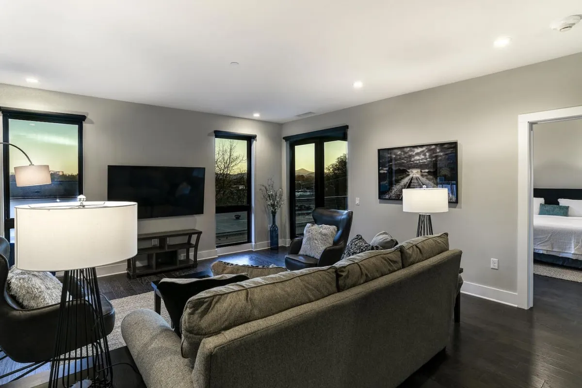 A stylish, comfortable living room with a plush sofa, modern lamp, and artwork, all oriented toward large windows. The space is designed for relaxation and entertainment, offering soft lighting and a welcoming mood for guests gathering after a day in the city.