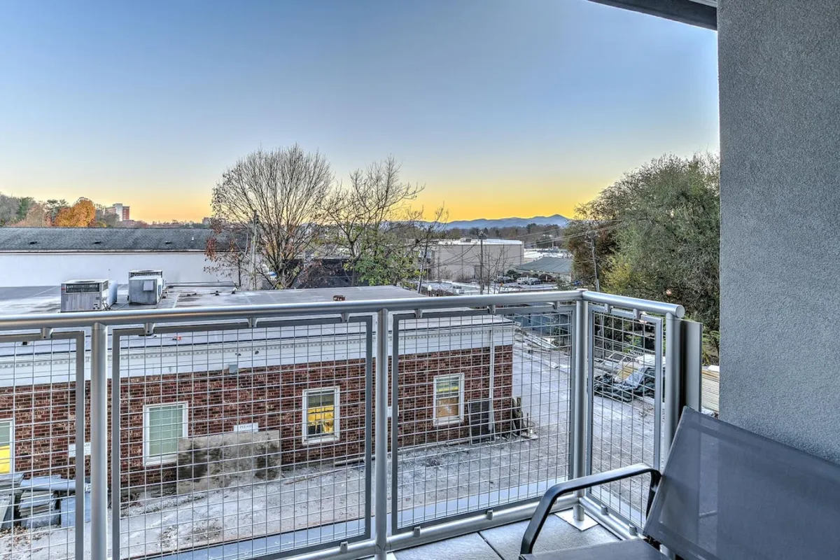 An inviting balcony overlooks tree-lined streets, distant mountain ranges, and the rooftops of downtown buildings. The scene evokes a serene transition from home to the vibrant urban life below, hinting at opportunities for relaxation or socializing before stepping out to explore the city.