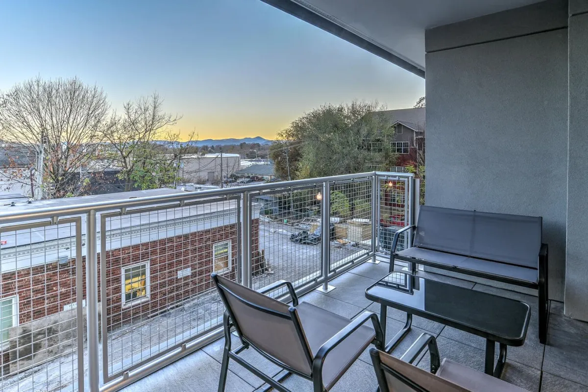 A serene balcony setting with comfortable seating and an expansive view of the Blue Ridge Mountains and Asheville’s urban landscape. The mood is tranquil, suitable for both morning relaxation and social gatherings, with the sunset’s glow hinting at memorable evenings.