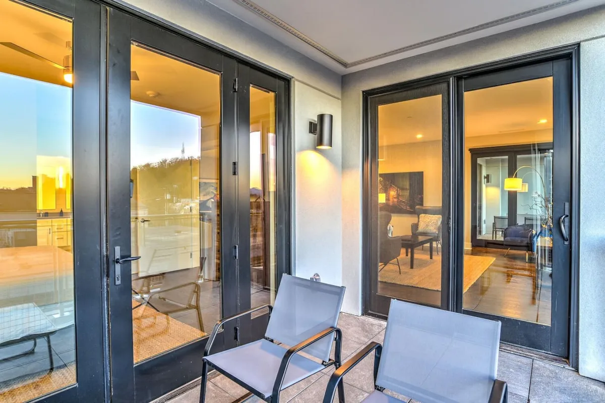 The image shows the exterior of the building featuring modern glass doors, outdoor seating, and warm interior lighting visible through the windows. The entryway is well-lit and inviting, suggesting a sense of security and urban sophistication.