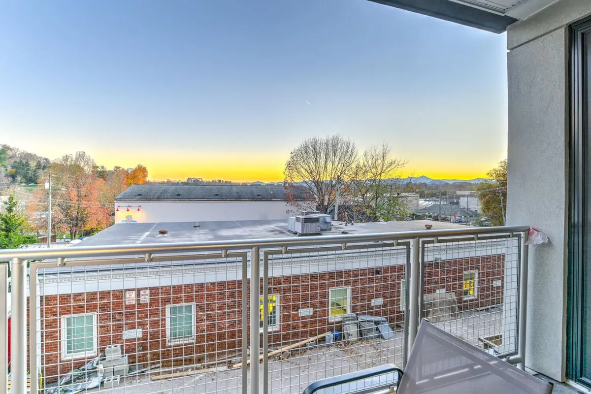 This image captures an exterior balcony scene featuring a sturdy railing, distant mountain views, and a vivid sunset sky. The atmosphere is peaceful, suggesting the condo’s connection to the outdoors and the scenic city surroundings. The sightline includes hints of neighboring rooftops, placing the viewer in a lively urban yet nature-kissed setting.