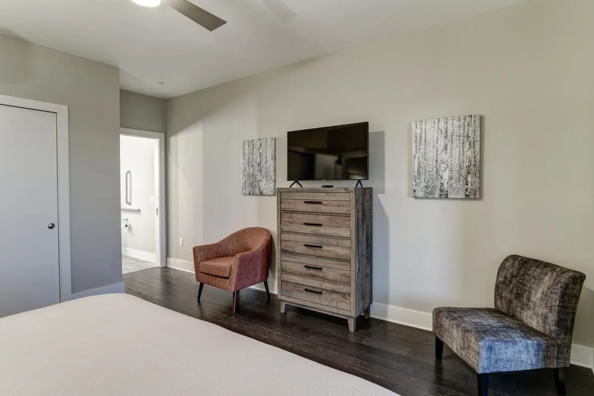 A tranquil bedroom with a neatly made bed, comfortable chairs, a television, a dressing table, and tasteful art pieces on the wall. The room features soft lighting and an inviting atmosphere that suggests a peaceful retreat after a day of exploring.