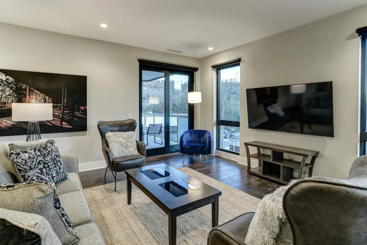 A bright, open living area featuring a comfortable sofa, coffee table, and striking artwork. Large windows frame views of the vibrant downtown Asheville scene, allowing abundant natural light to fill the space. The arrangement invites guests to relax, socialize, or take in the city atmosphere from a cozy vantage point.