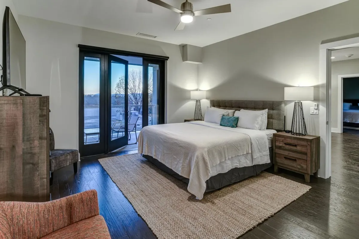 A tranquil king bedroom featuring a plush king-sized bed, soft linens, nightstands with lamps on either side, seating area, and large balcony doors that open to fresh mountain views. The room is beautifully lit with natural light, and the decor exudes understated luxury and comfort.