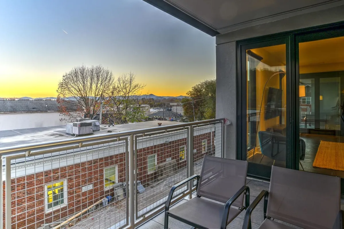 A peaceful balcony scene featuring a sturdy railing overlooking majestic mountains in the distance. Tree branches gently frame the view, and the interior room is subtly visible through the glass doors, suggesting seamless indoor-outdoor living. The space is ready for quiet mornings or celebratory evenings.