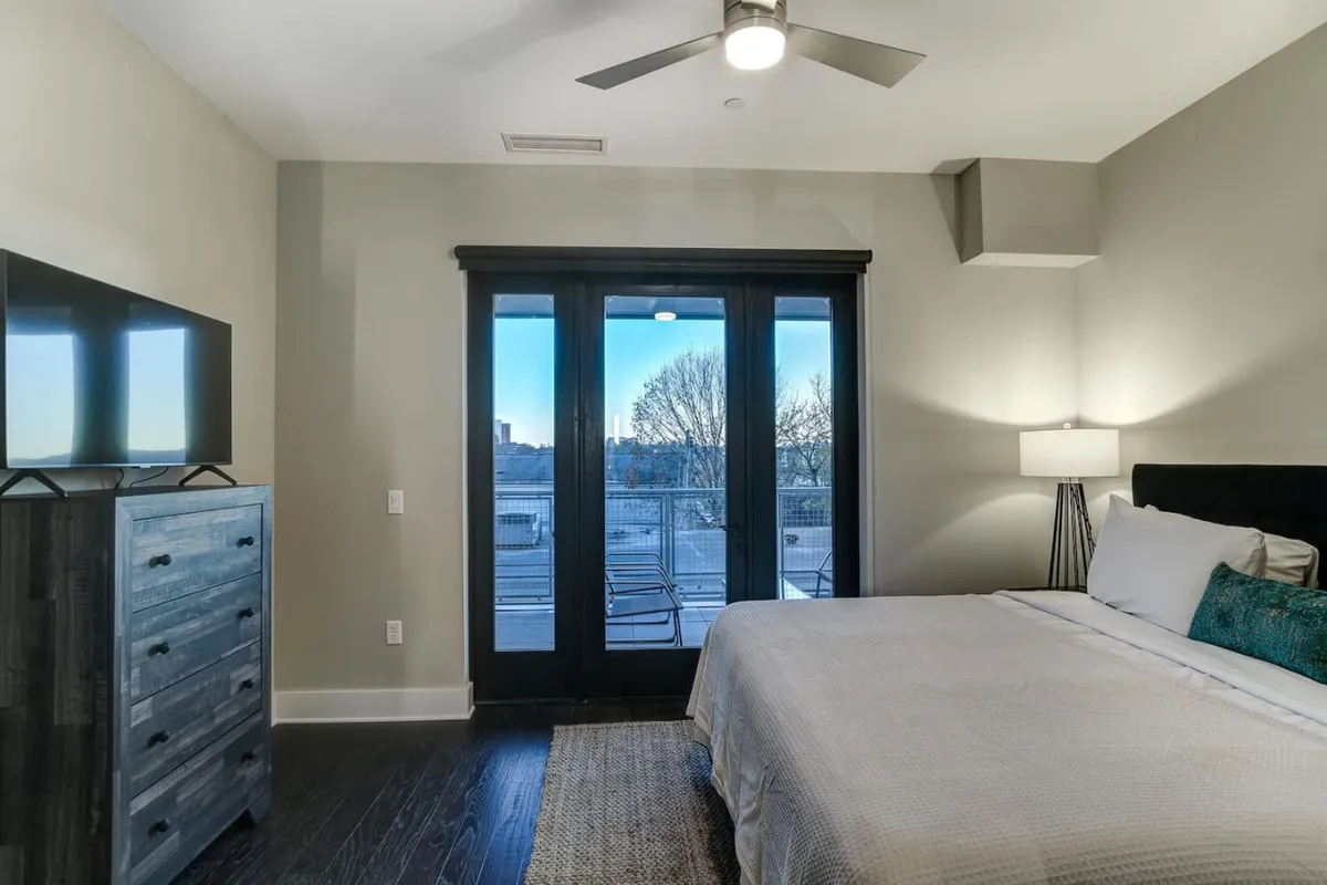 An inviting bedroom featuring a plush bed, large windows, a dresser, and a lamp. Natural light pours in, hinting at the proximity of a balcony just beyond the frame, and inviting morning routines with beautiful views.